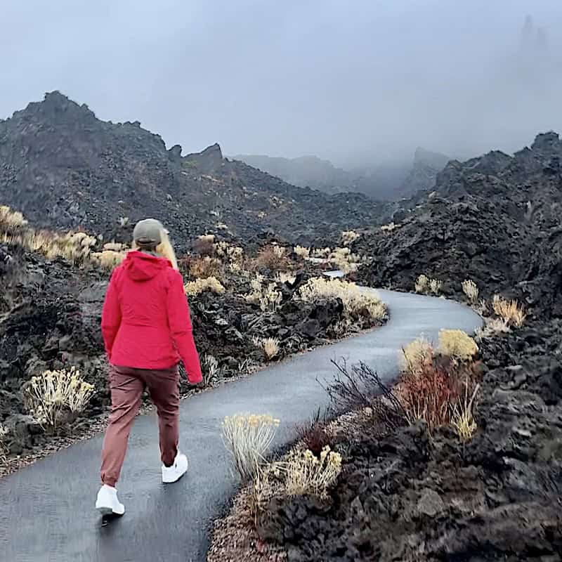 a woman wearing a red rain jacket walks on a paved path through a black cinder lava flow in Oregon